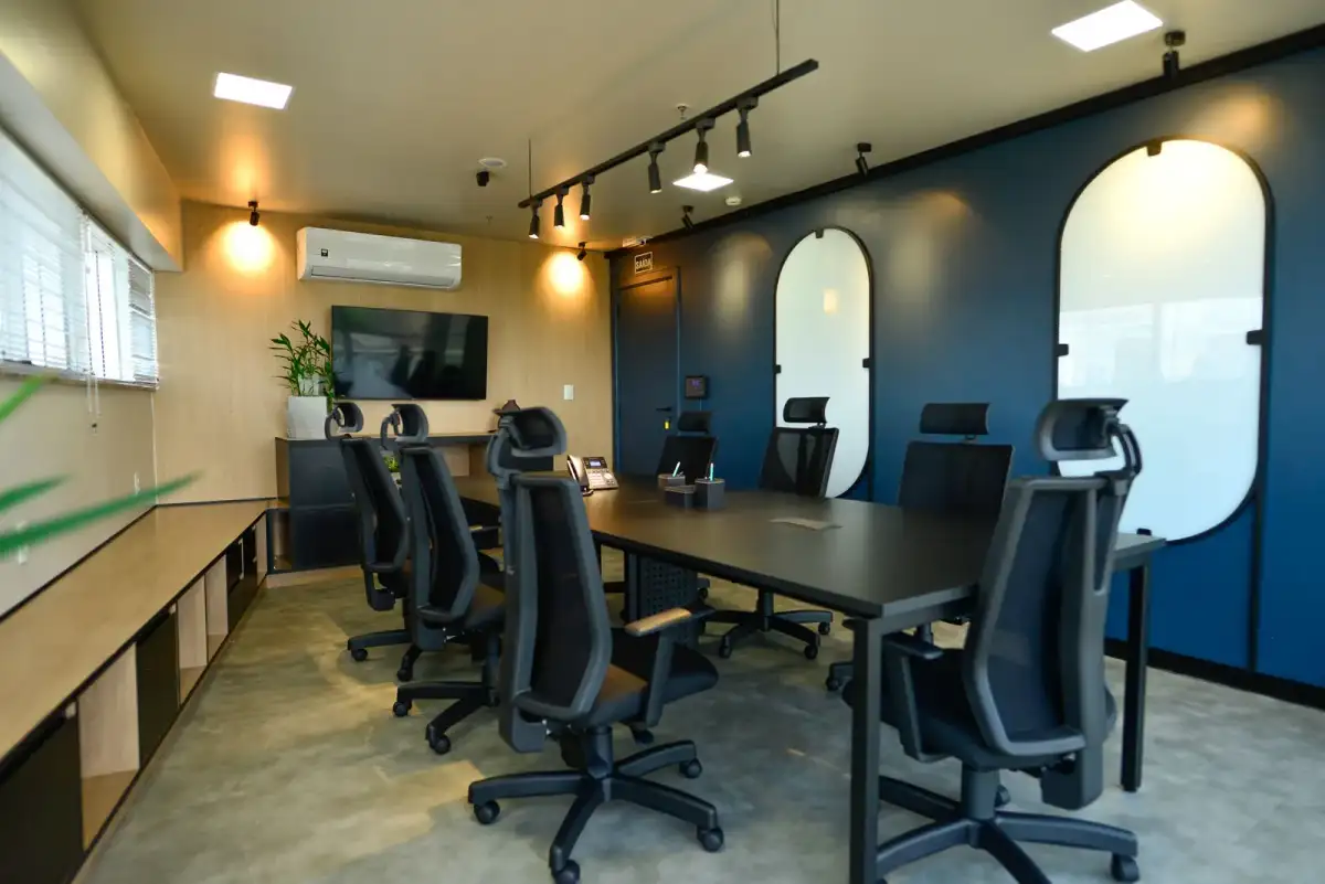 Sala de reunião com mesa preta, cadeiras de tela, parede azul com portas em arco e tela na parede