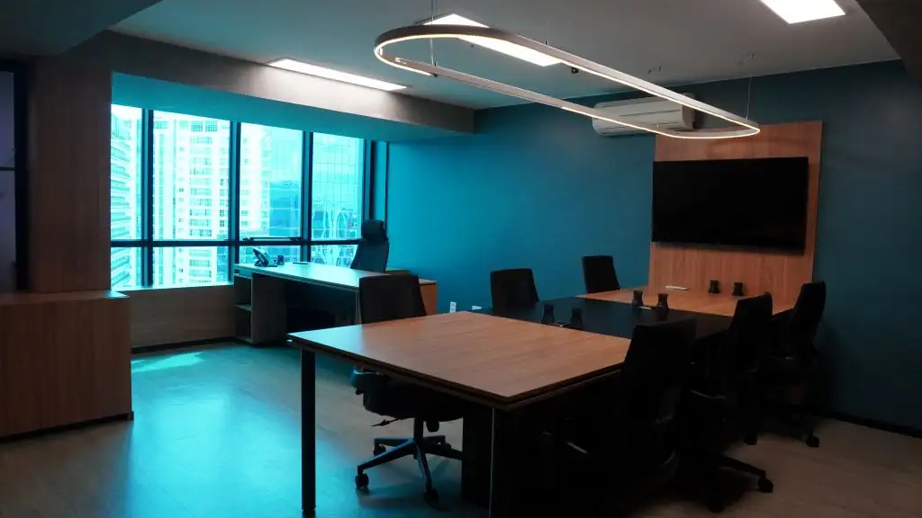 Sala de equipe com mesas em madeira, luminária circular e janelas com vista para os prédios