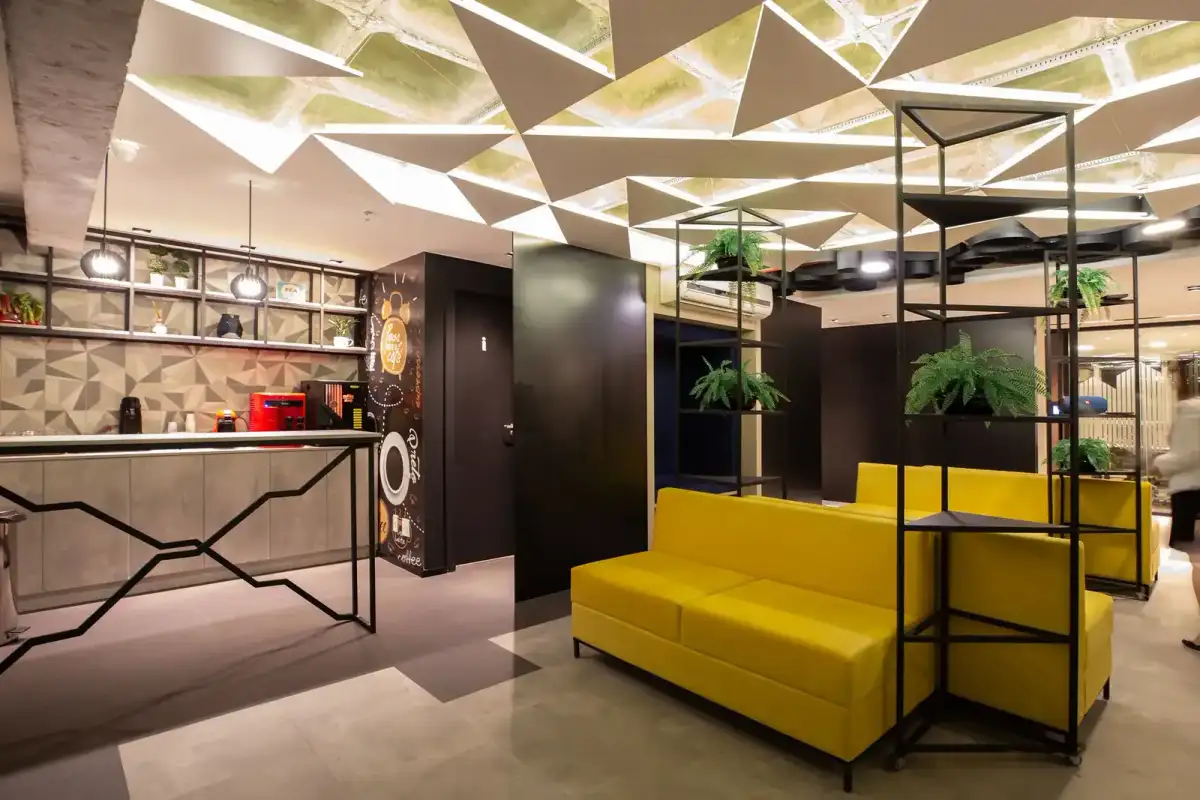 Lounge da unidade Prime Business com sofás amarelos, estantes de ferro com plantas e teto geométrico