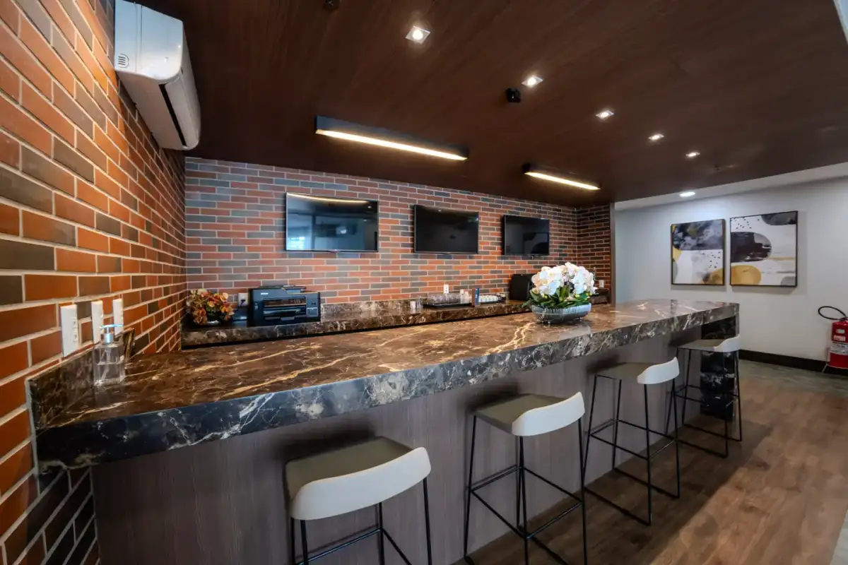 Café da unidade Nasa Business Style com bancada de mármore, banquetas, parede de tijolos e teto de madeira