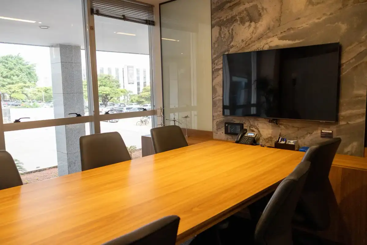 Escritório privativo da unidade Prime Business com mesa de reunião, painel de pedra, tela e janela para a cidade