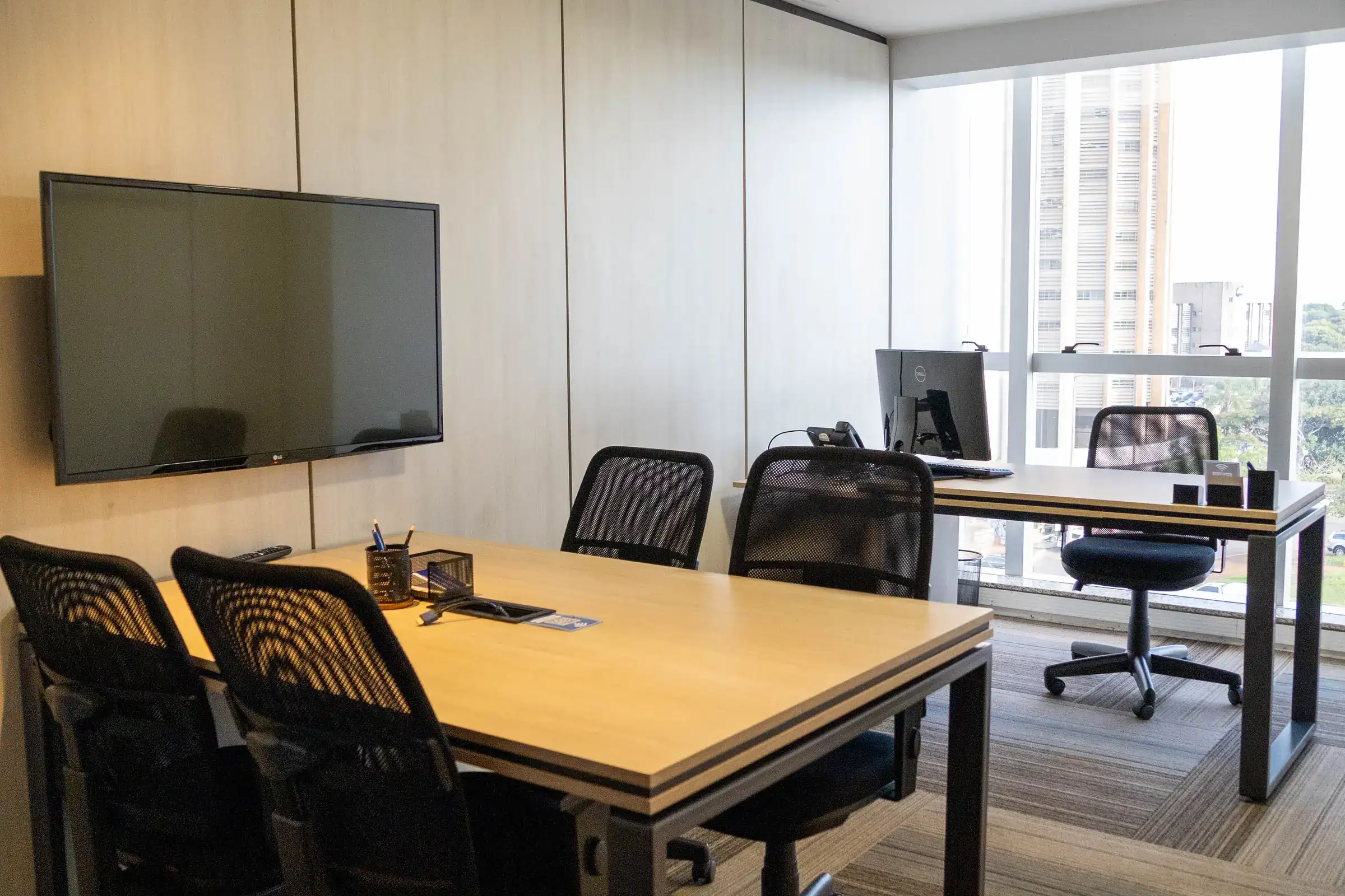 Escritório privativo mobiliado da unidade Prime Business com mesa de trabalho, mesa de reunião, tela e janela com vista para a cidade